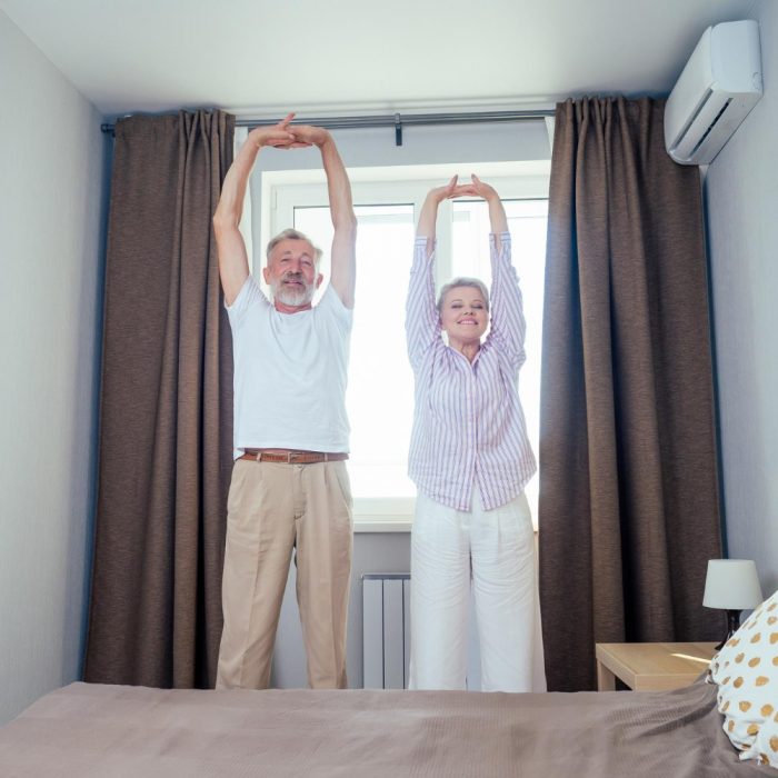 blonde-short-haircut-woman-nightwear-elderly-man-doing-stretching-handsspine-day-light-bedroom-apartment-big-window-with-brown-curtains-air-conditioning-background